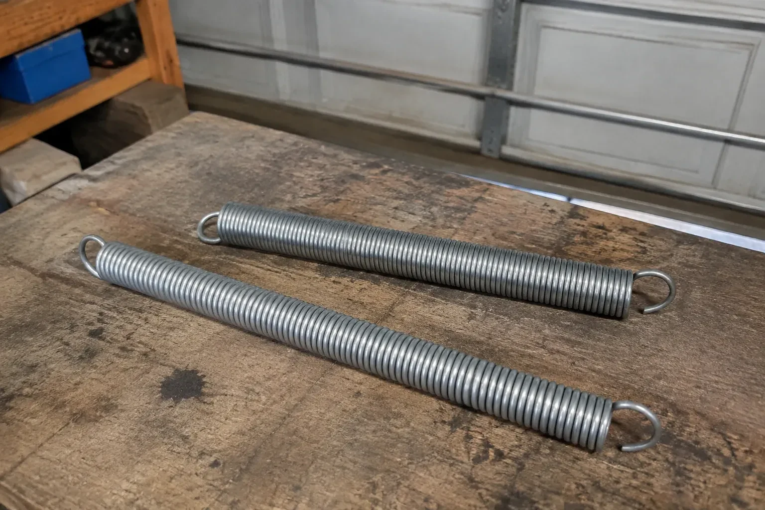 Two detached extension springs with hooked ends on a garage bench. — Extension Spring Replacement, Centre Wellington