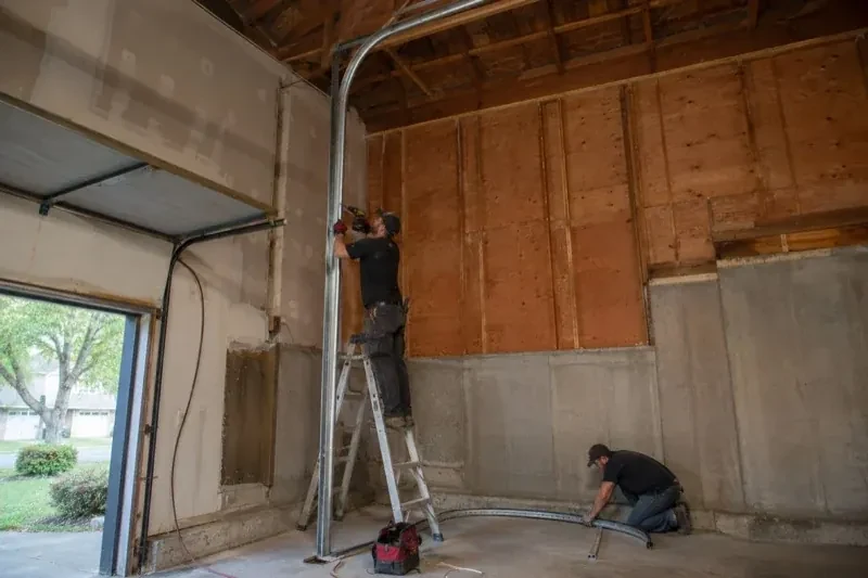 One person on a stepladder working beside a tall curved garage-door track while another kneels beside loose track sections in an unfinished garage. — High-Lift Garage Door Conversion, Centre Wellington